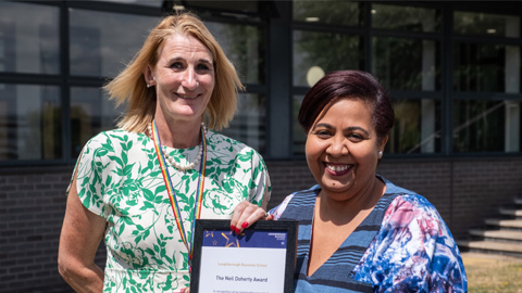 Two people standing in front of a building with large windows; one holds a framed certificate or award.
